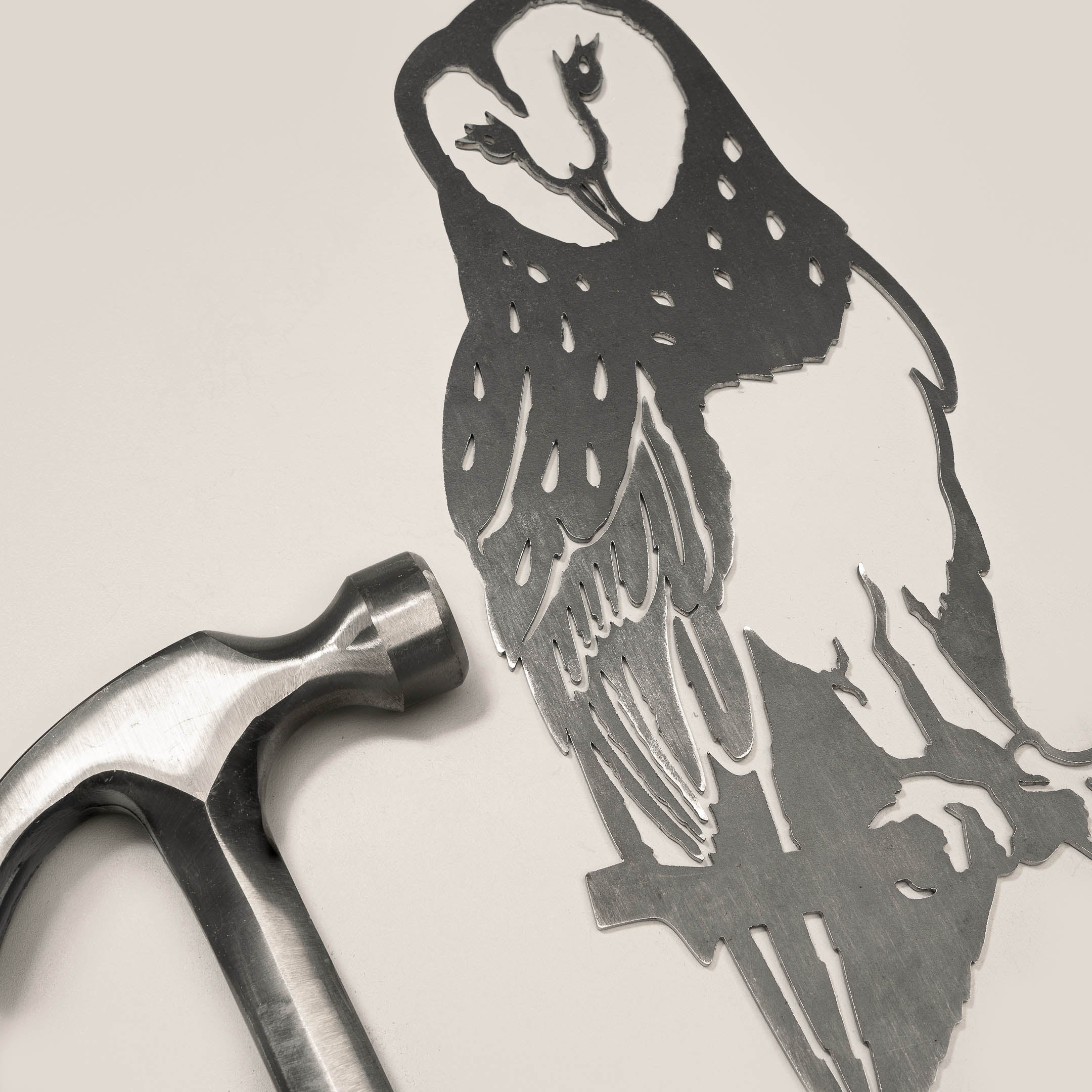 A silver hammer lies next to the Metalbird UK Barn Owl, a detailed corten steel owl silhouette with unique feather patterns, both arranged on a light beige surface.