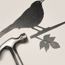The Blackbird by Metalbird UK features a metal bird silhouette perched on a leafy branch beside a hammer, making unique yard art or a thoughtful gift, all shown against a light background.