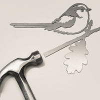 The Metalbird UK Long-tailed Tit features a flat metal bird silhouette perched on an oak branch, with a hammer included for easy installation—perfect as eye-catching yard art or a unique gift.