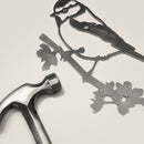 A metal hammer lies beside the Metalbird UK Blue Tit garden art cutout, featuring a bird on a branch with flowers, all set against a plain light background.