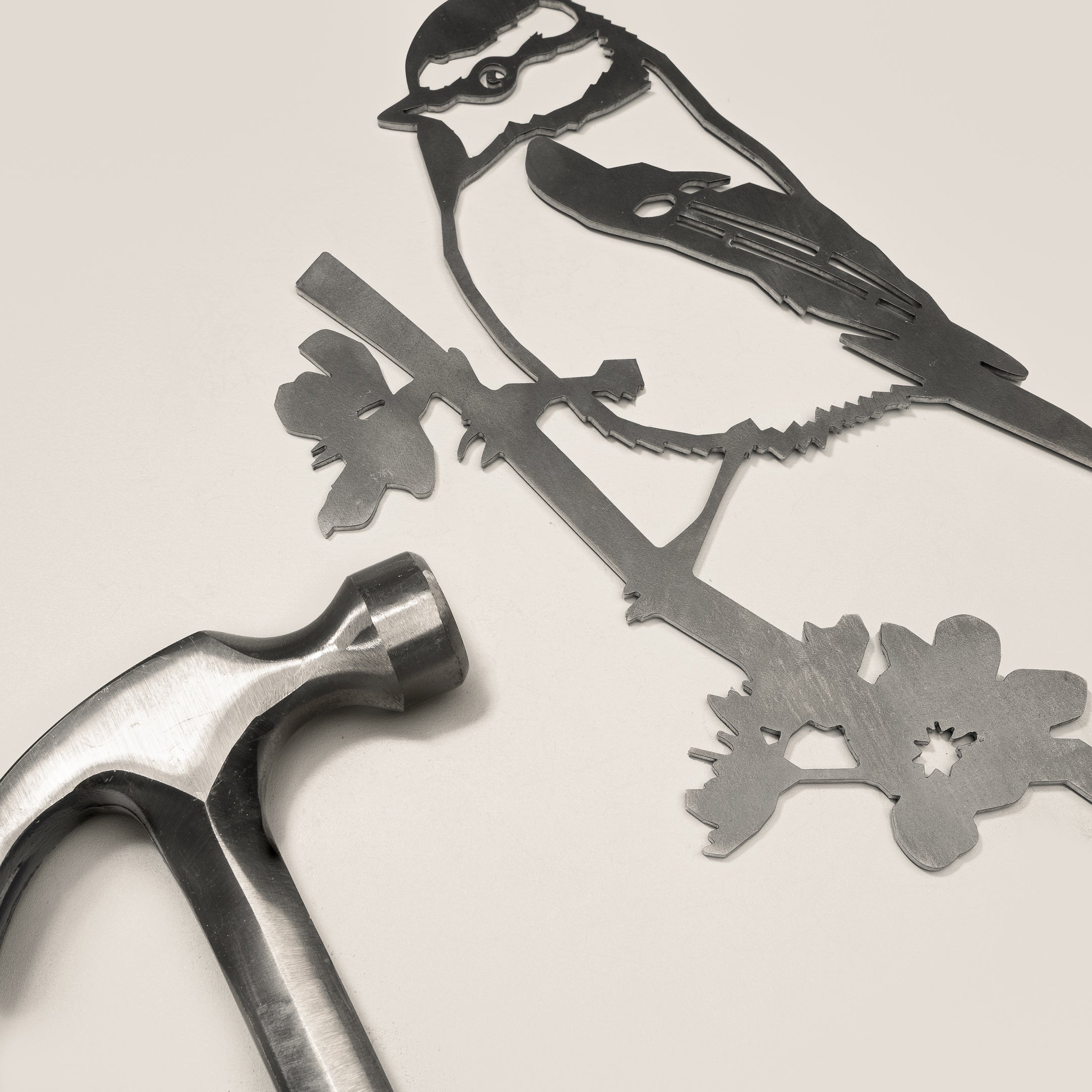 A metal hammer lies beside the Metalbird UK Blue Tit garden art cutout, featuring a bird on a branch with flowers, all set against a plain light background.