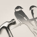 A metal hammer sits next to the Metalbird UK "Pair of Swallows" flat metal silhouette cutout—two birds on a branch—ideal as garden art, all displayed on a light-colored surface.