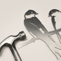 A metal hammer sits next to the Metalbird UK "Pair of Swallows" flat metal silhouette cutout—two birds on a branch—ideal as garden art, all displayed on a light-colored surface.