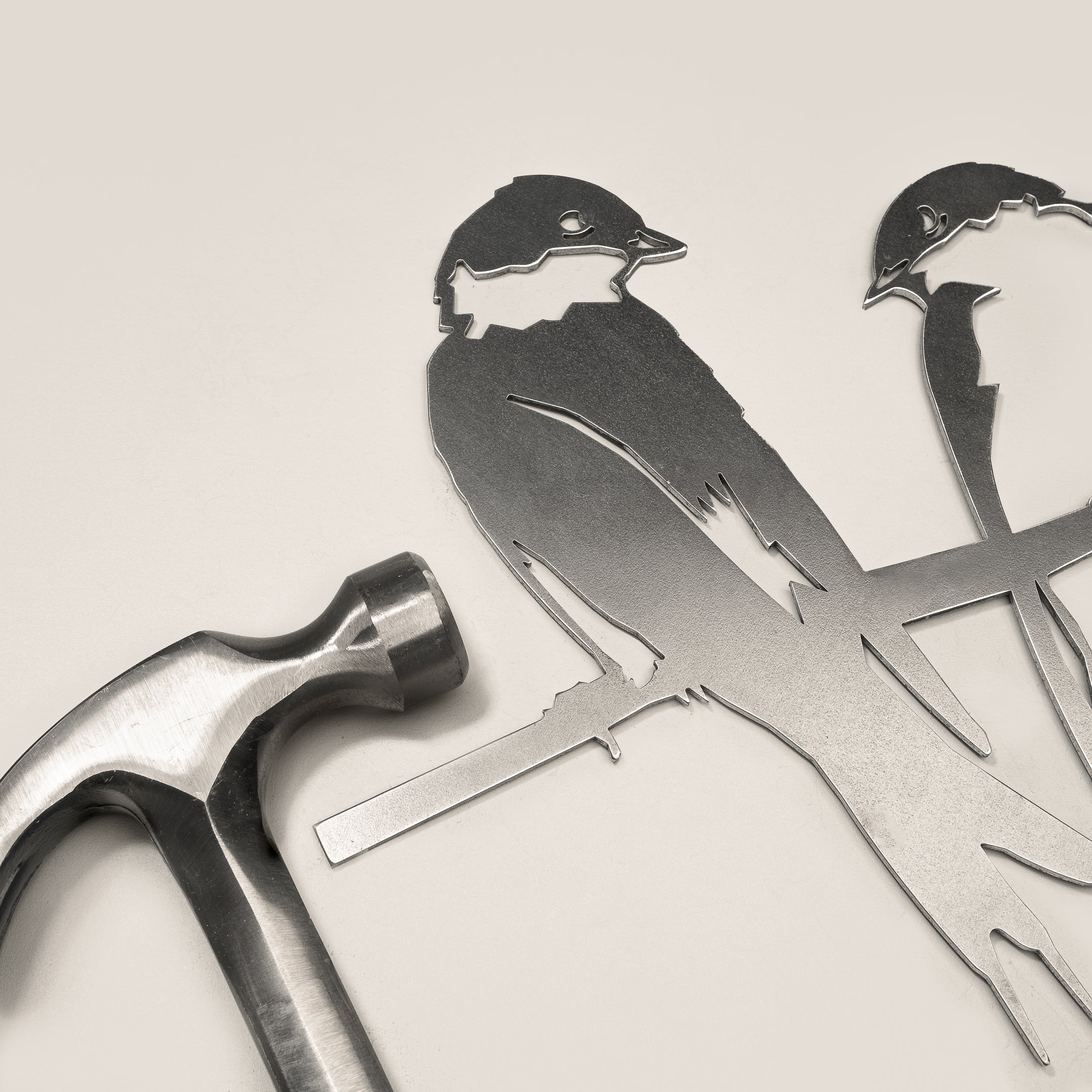 A metal hammer sits next to the Metalbird UK "Pair of Swallows" flat metal silhouette cutout—two birds on a branch—ideal as garden art, all displayed on a light-colored surface.