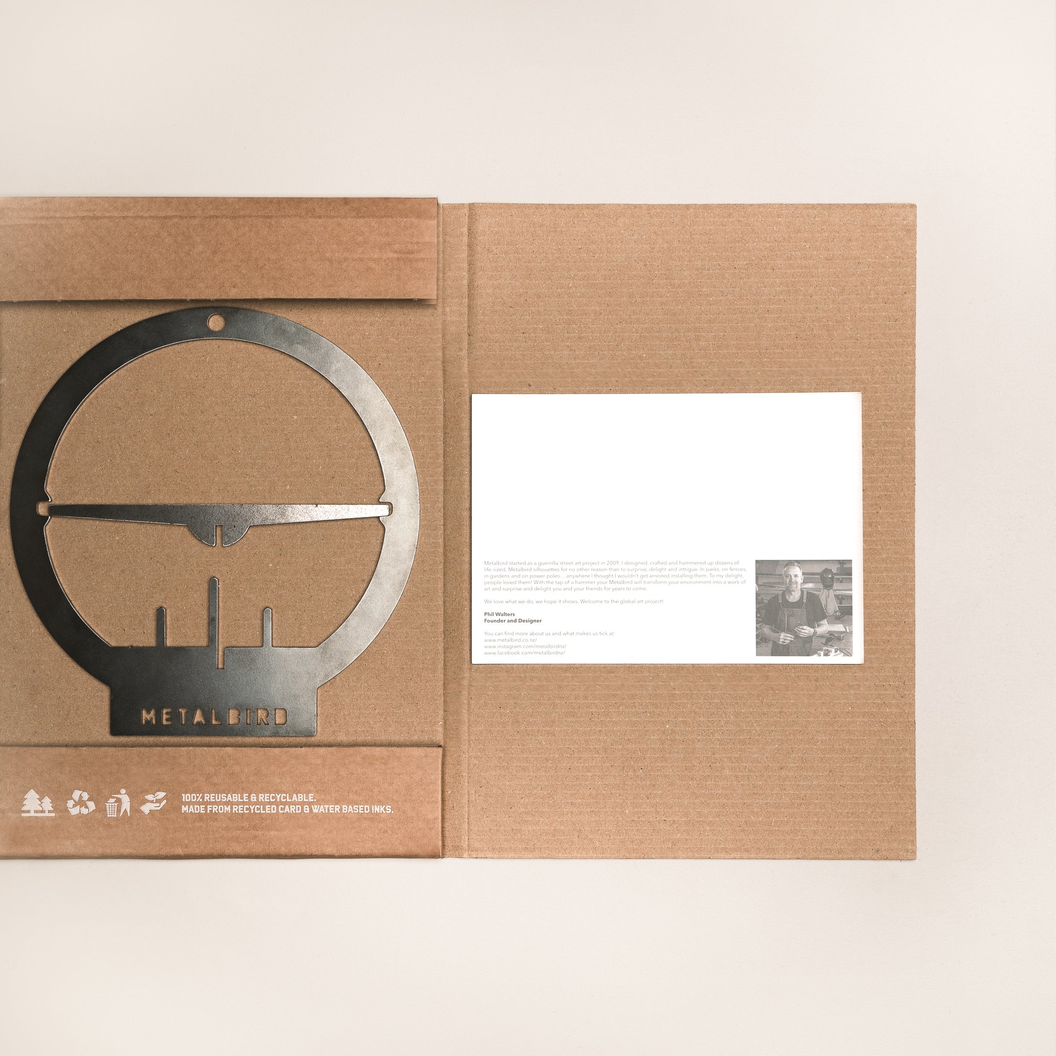 The Metalbird UK Hanging Feeder is a flat, circular corten steel art piece with a bird and tree branches, packaged in a brown folder with a white card and photo—making it a unique gift.