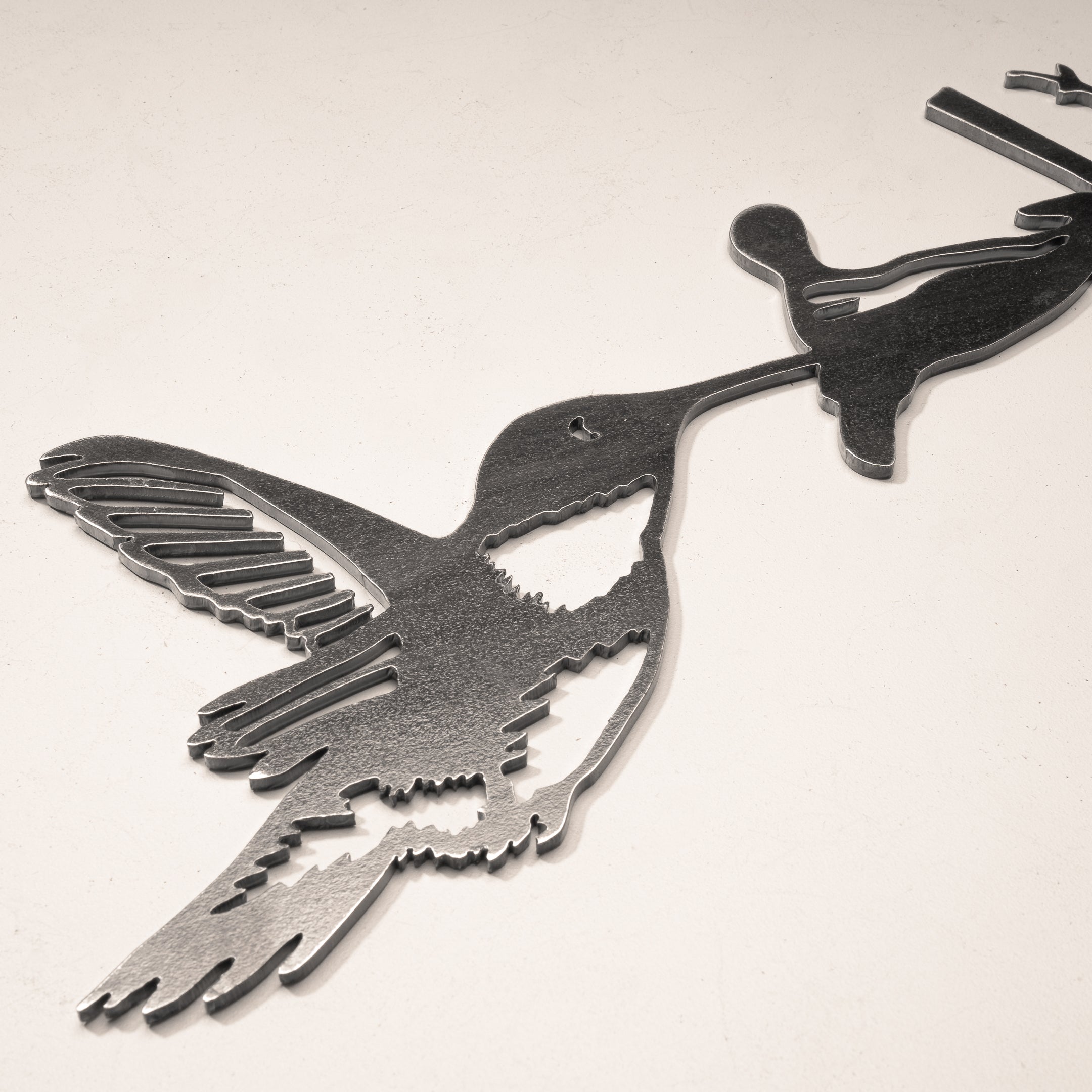 The Hummingbird by Metalbird UK is a metal wall art piece depicting a hummingbird drinking nectar, ideal as garden art and stunning when displayed on a light-colored textured surface.