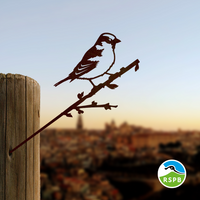A metal House Sparrow cutout by Metalbird UK perches on a wooden post, standing out as striking yard art against a blurred cityscape and sunset—an ideal gift for nature lovers.
