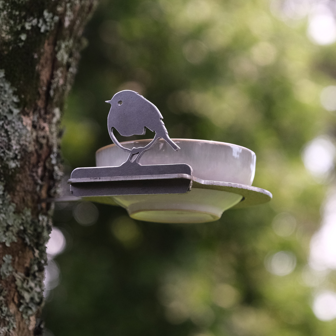 The Bowl Bird Feeder by Metalbird UK features a white ceramic cup on a metal saucer with a bird silhouette, mounted on a tree trunk—a charming garden art piece set against green foliage.