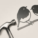 The Metalbird UK Robin Pair features two flat, corten steel bird silhouettes perched on a branch, displayed beside a metal hammer against a light background.