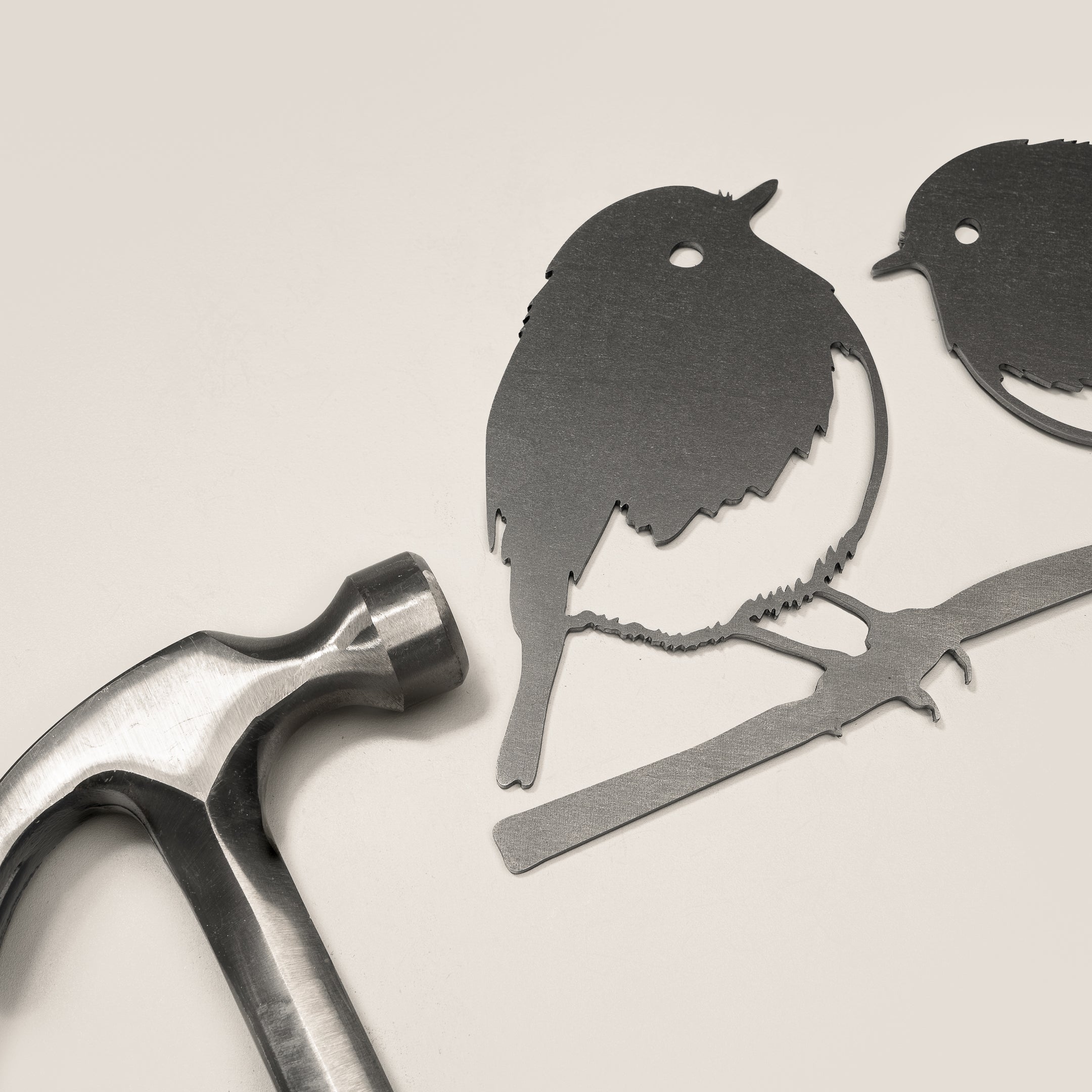 The Metalbird UK Robin Pair features two flat, corten steel bird silhouettes perched on a branch, displayed beside a metal hammer against a light background.