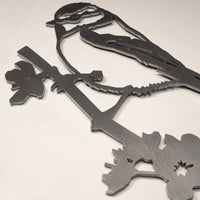The Metalbird UK Blue Tit is a metal silhouette of a bird perched on a branch with flowers, making a charming piece of garden art against any light, neutral background.