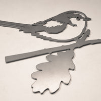 The Long-tailed Tit by Metalbird UK is a metallic, geometric silhouette of a bird on a branch with a large leaf, crafted from corten steel. Its clean lines and striking design make it standout yard art for any light-colored surface.