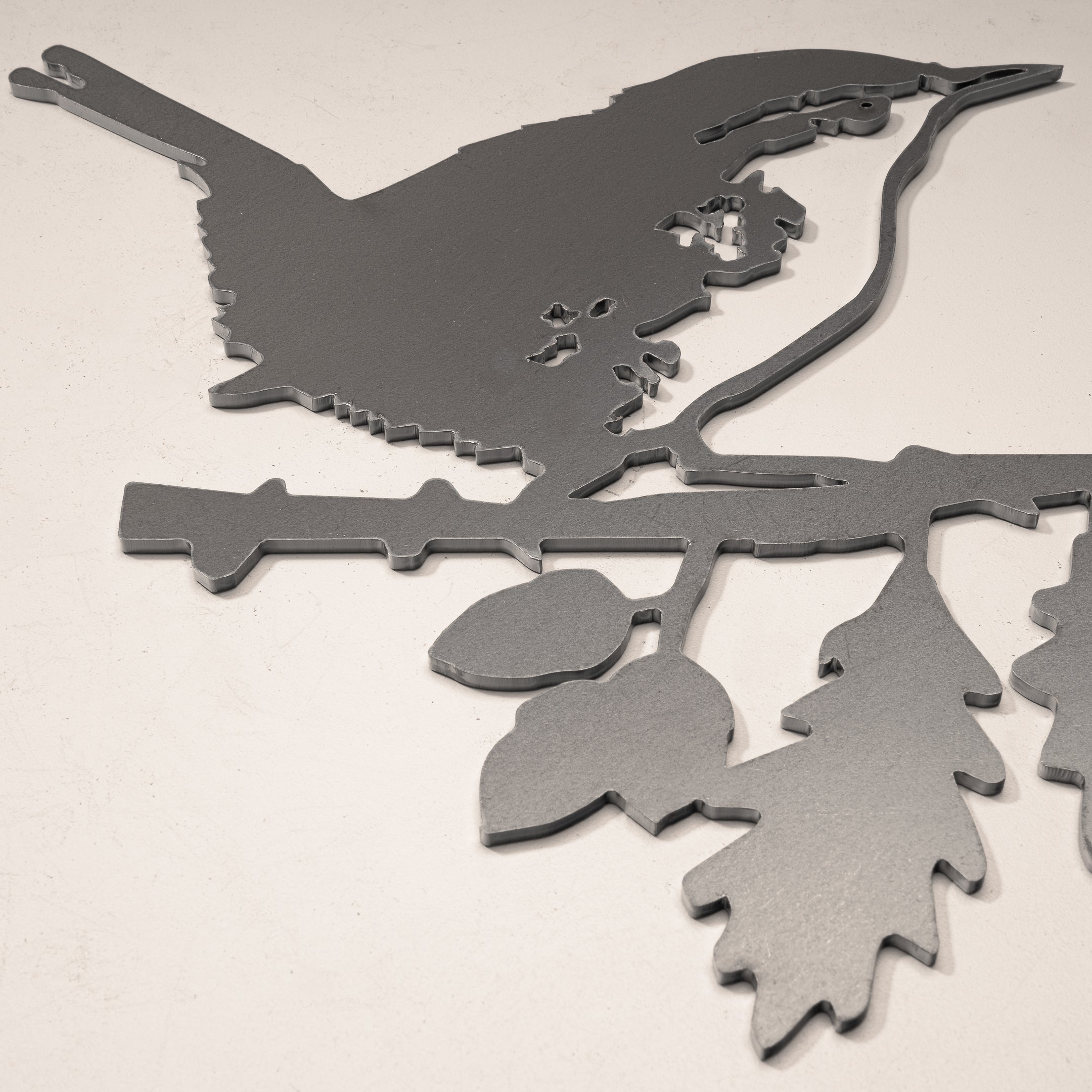 The Metalbird UK Wren is a minimalist, flat corten steel silhouette showing a wren perched on a leafy branch in profile, displayed on a light-colored surface.