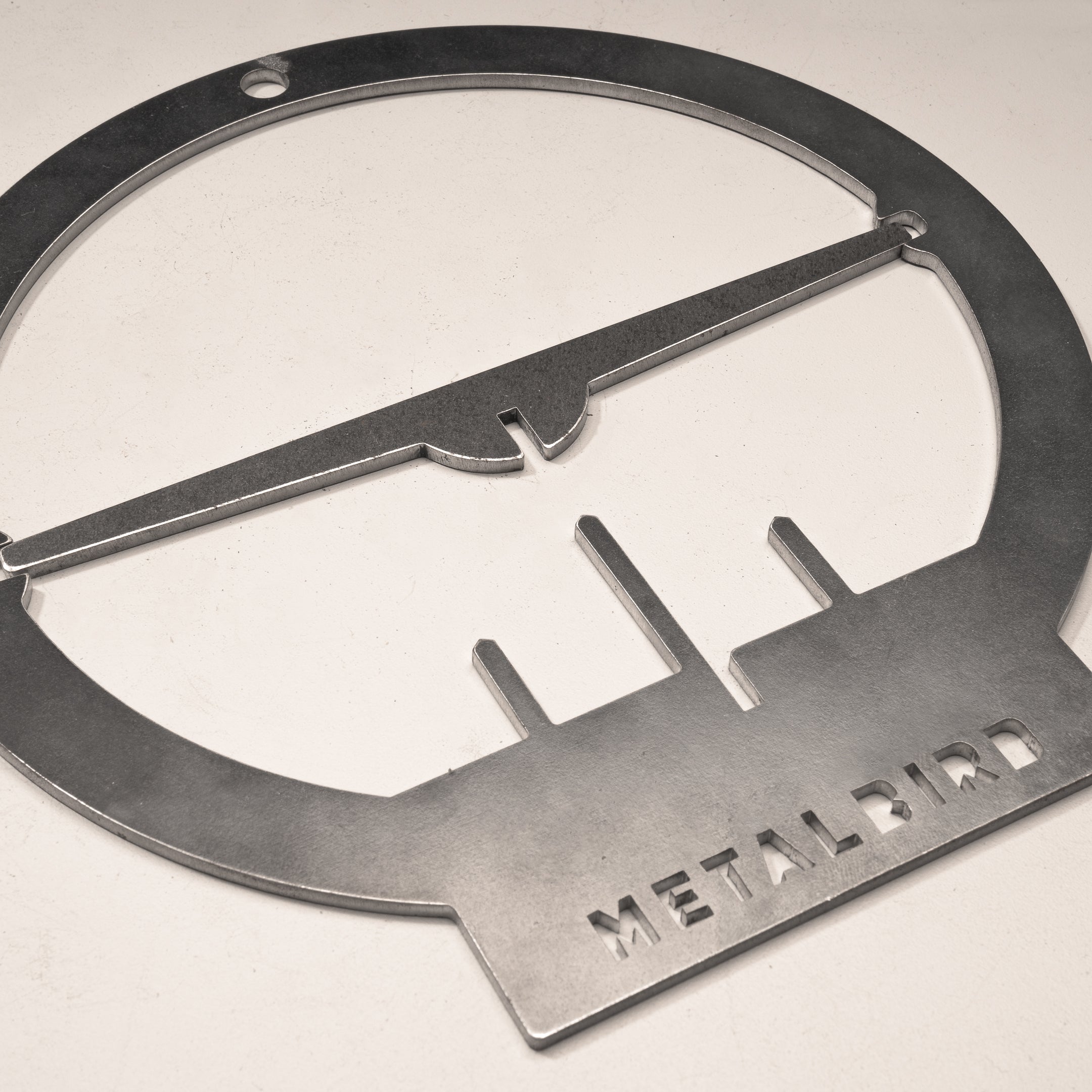 A brushed metal Hanging Feeder by Metalbird UK features a minimalist bird perched on a branch within a circular frame, with "METALBIRD" cut out at the base—an eye-catching sculpture ideal for unique garden art.