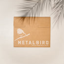 Brown cardboard envelope with a white silhouette of a raven on a branch and the text "Metalbird UK Proudly Made Here in the UK." A shadow of palm leaves falls across the top right corner. Perfect for raven art lovers.
