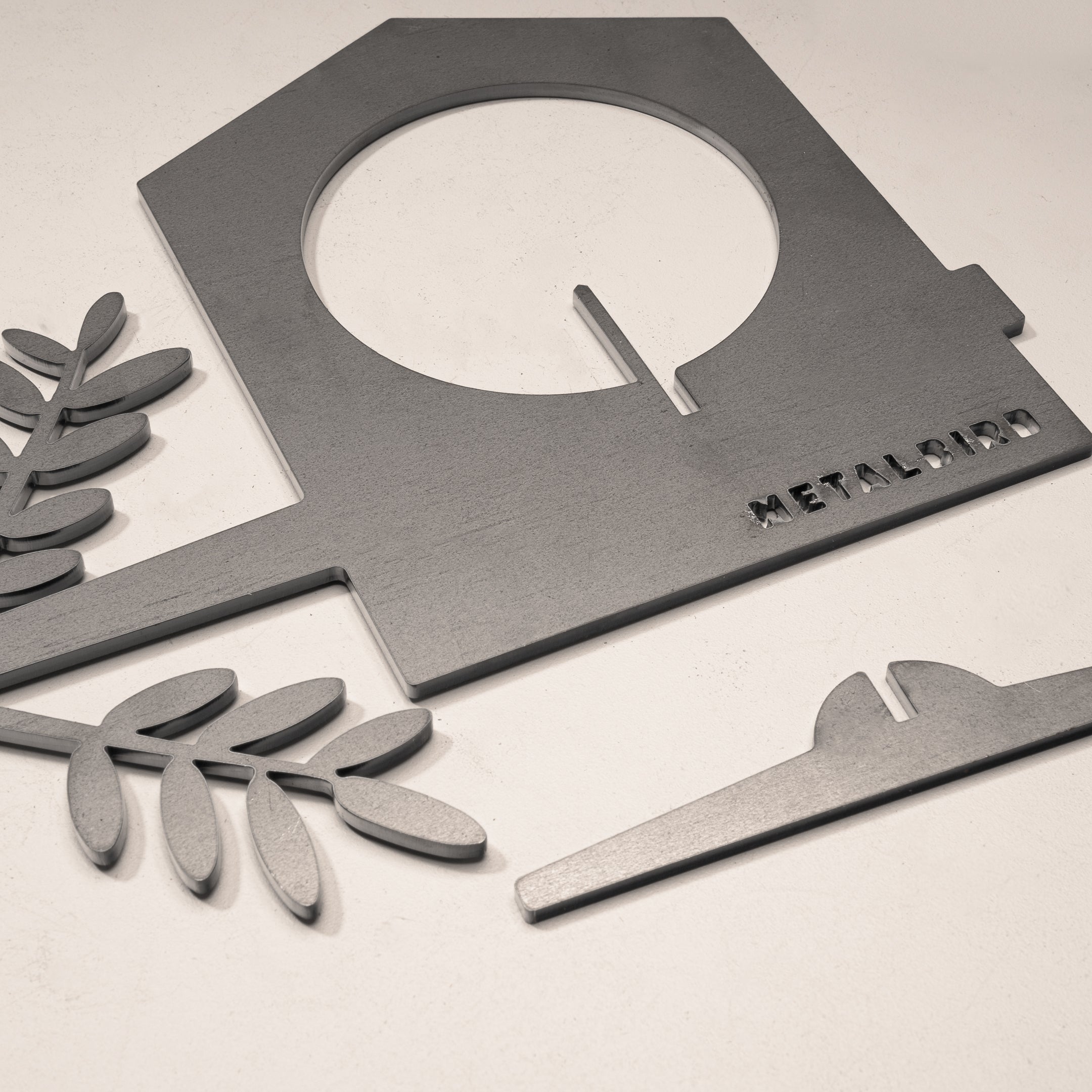 A gray corten steel Bird Feeder by Metalbird UK, featuring a house-shaped frame with circular cutout, two leaf-shaped parts, and a flat rectangle, is arranged on a light surface. Each feeder is engraved with the Metalbird brand.