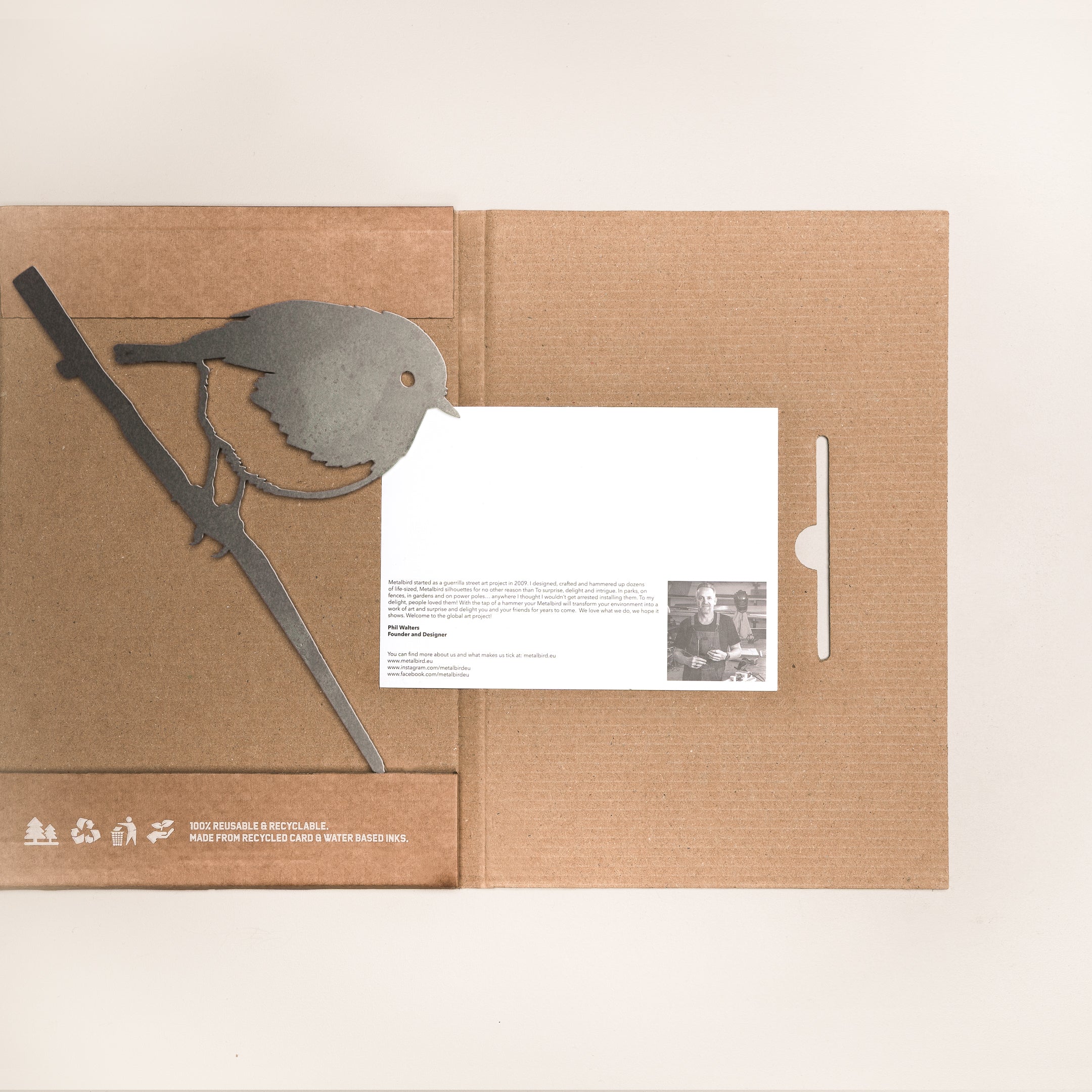 The Robin by Metalbird UK features a metal bird silhouette perched on a branch, an illustrated cardboard folder with recycling symbols, and an info card—all displayed on a light background—ideal as decorative yard art.