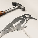 A metal hammer with a wooden handle sits beside Metalbird UK’s Great Spotted Woodpecker—a flat corten steel garden art piece—on a light surface.