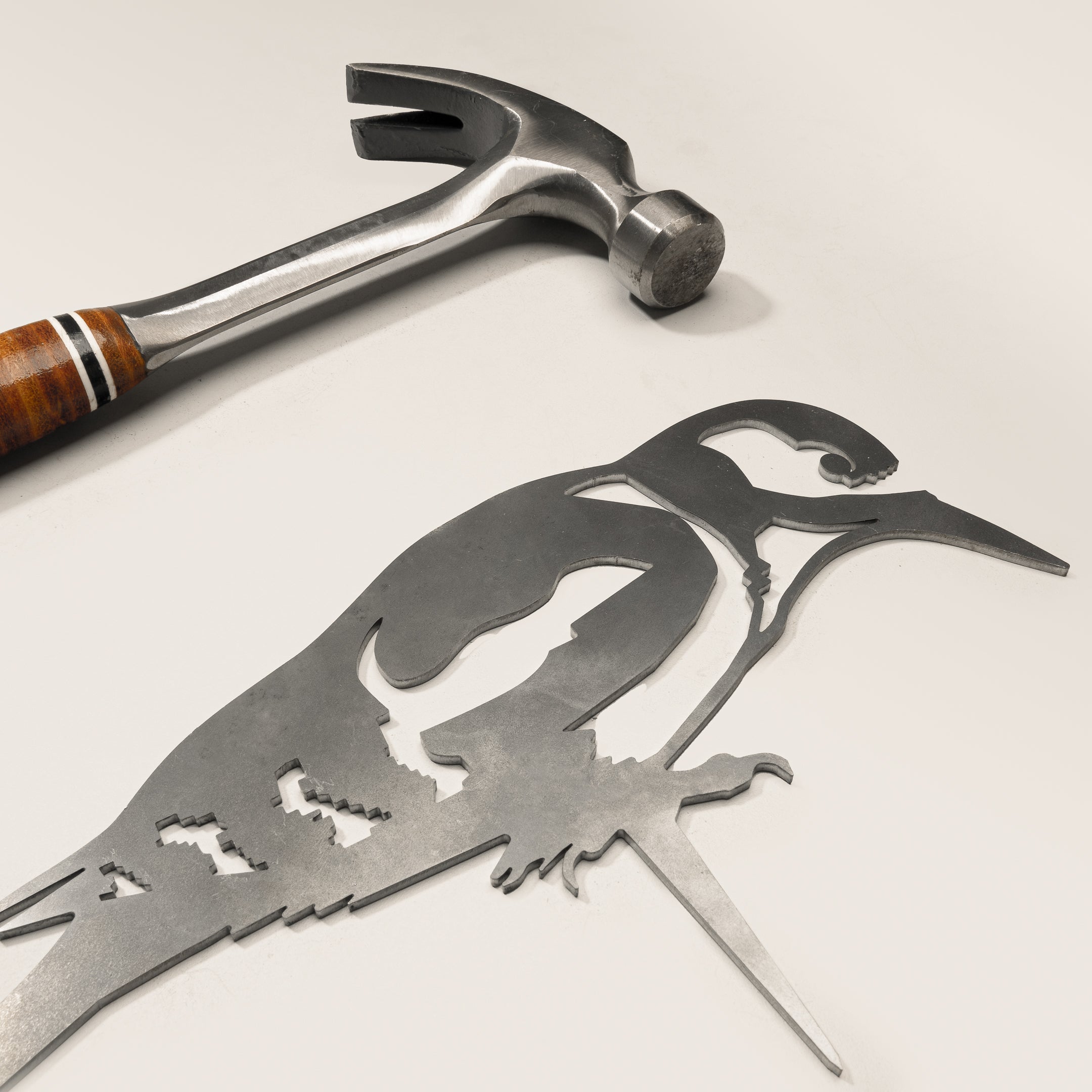 A metal hammer with a wooden handle sits beside Metalbird UK’s Great Spotted Woodpecker—a flat corten steel garden art piece—on a light surface.