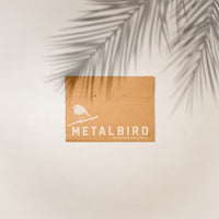 A brown cardboard envelope with "Metalbird UK" and a bird illustration in white holds the Hanging Feeder, perfect for gifting unique yard art. It rests on a light surface, with palm leaf shadows above.