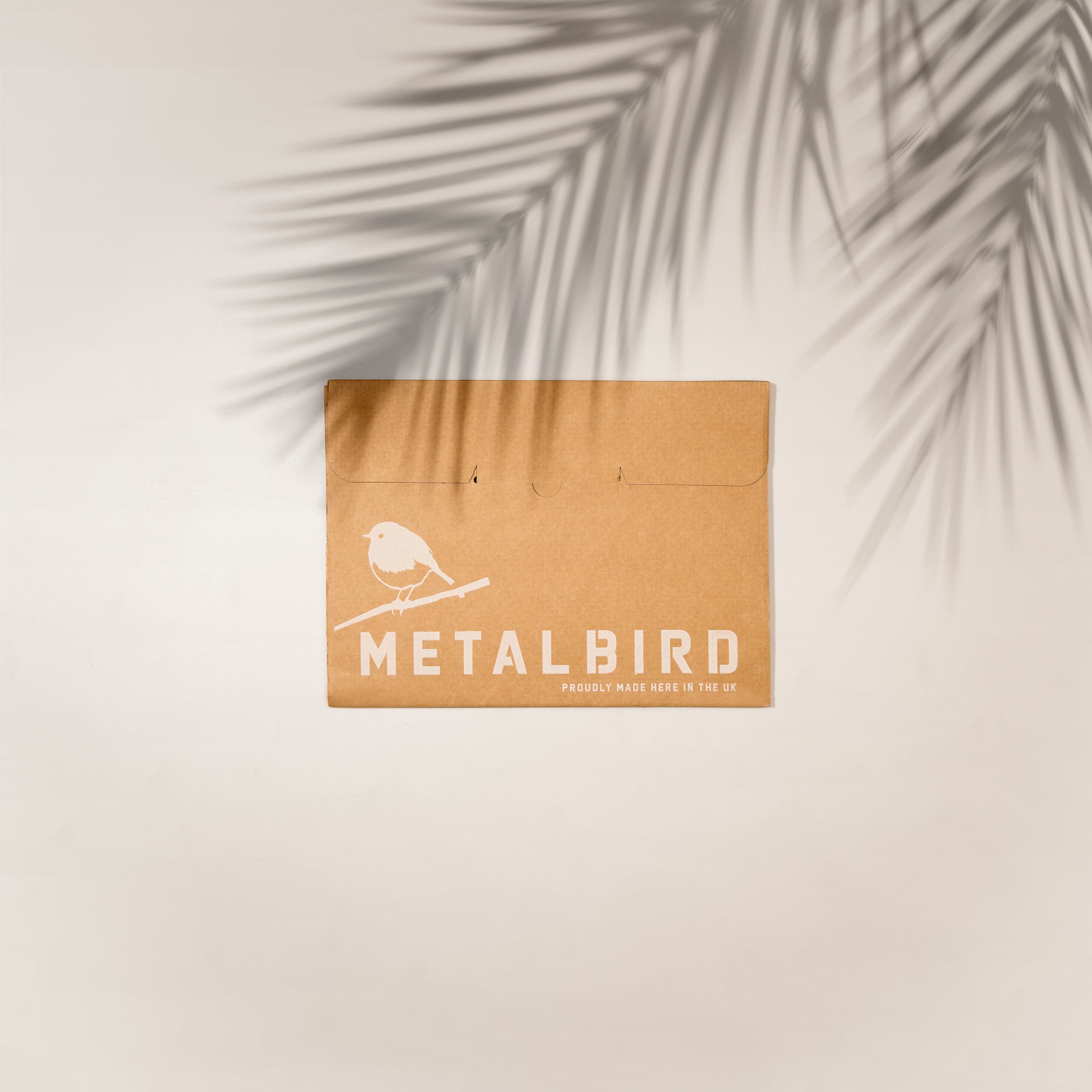 A brown cardboard envelope with "Metalbird UK" and a bird illustration in white holds the Hanging Feeder, perfect for gifting unique yard art. It rests on a light surface, with palm leaf shadows above.