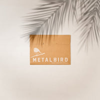 A brown envelope with a white bird silhouette and "METALBIRD" text, from Metalbird UK’s Bowl Bird Feeder, rests against a light background. Leafy plant shadows create a charming scene that echoes metal yard art.