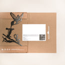 A flat cardboard package from Metalbird UK with a metallic Hummingbird and flower silhouette cutout, partially open to reveal a white card inside. Eco-friendly icons and recyclable text make it ideal for gifting unique yard art.