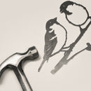 A metal hammer rests beside Metalbird UK's Pair of Sparrows, a delicate corten steel silhouette of two birds on branches, set against a light, neutral background.
