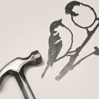 A metal hammer rests beside Metalbird UK's Pair of Sparrows, a delicate corten steel silhouette of two birds on branches, set against a light, neutral background.