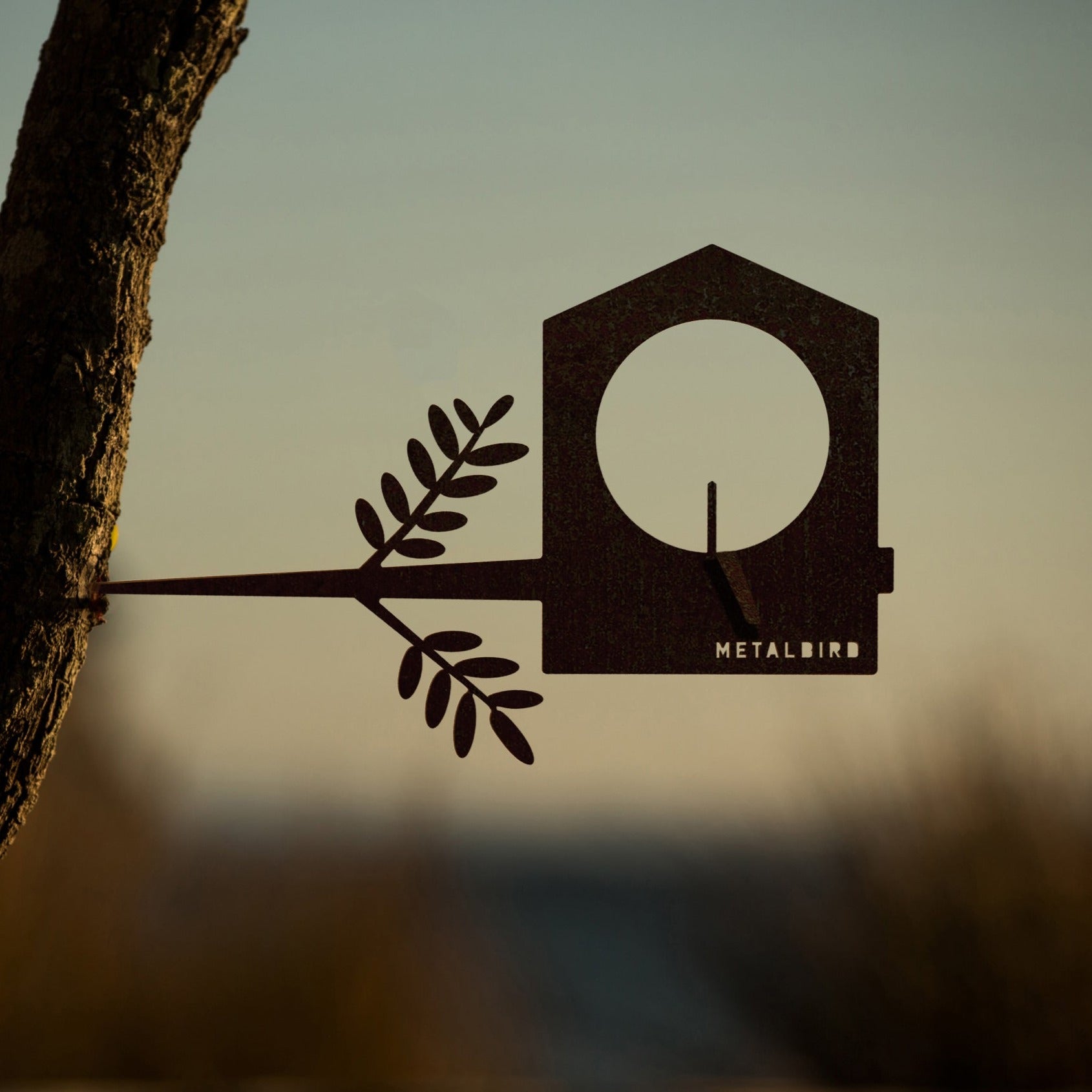The Metalbird UK Bird Feeder features a metal bird silhouette with branch detail, attached to a tree trunk against a sunset, making it unique yard art and a charming gift for nature lovers. The brand name is visible on the feeder.
