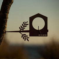 The Metalbird UK Bird Feeder features a metal bird silhouette with branch detail, attached to a tree trunk against a sunset, making it unique yard art and a charming gift for nature lovers. The brand name is visible on the feeder.