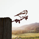 The Metalbird UK Blue Tit features a corten steel bird perched on a branch, mounted on a weathered wooden post against a blurred sunset landscape of rolling hills and fields.