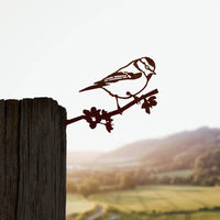 The Metalbird UK Blue Tit features a corten steel bird perched on a branch, mounted on a weathered wooden post against a blurred sunset landscape of rolling hills and fields.
