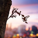 The Hummingbird by Metalbird UK, a corten steel silhouette of a hummingbird and flower, is mounted on a tree trunk with a blurred cityscape and vibrant sunset sky in the background.