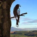 The Metalbird UK Harris Hawk is a corten steel silhouette of a bird on a branch, mounted on a tree with rolling hills and blue sky in the background—a striking piece of yard art.