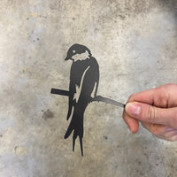 A hand holds the Metalbird UK Mini Swallow, a flat black metal silhouette of a bird on a branch—ideal as unique garden art or a charming gift—shown against a gray concrete background.