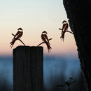 The Baby Bird Bundle - Swallow Trio by Metalbird UK features three metal swallows—two perched on a wooden post, one on a tree—creating striking yard art silhouettes against vibrant sunset skies.