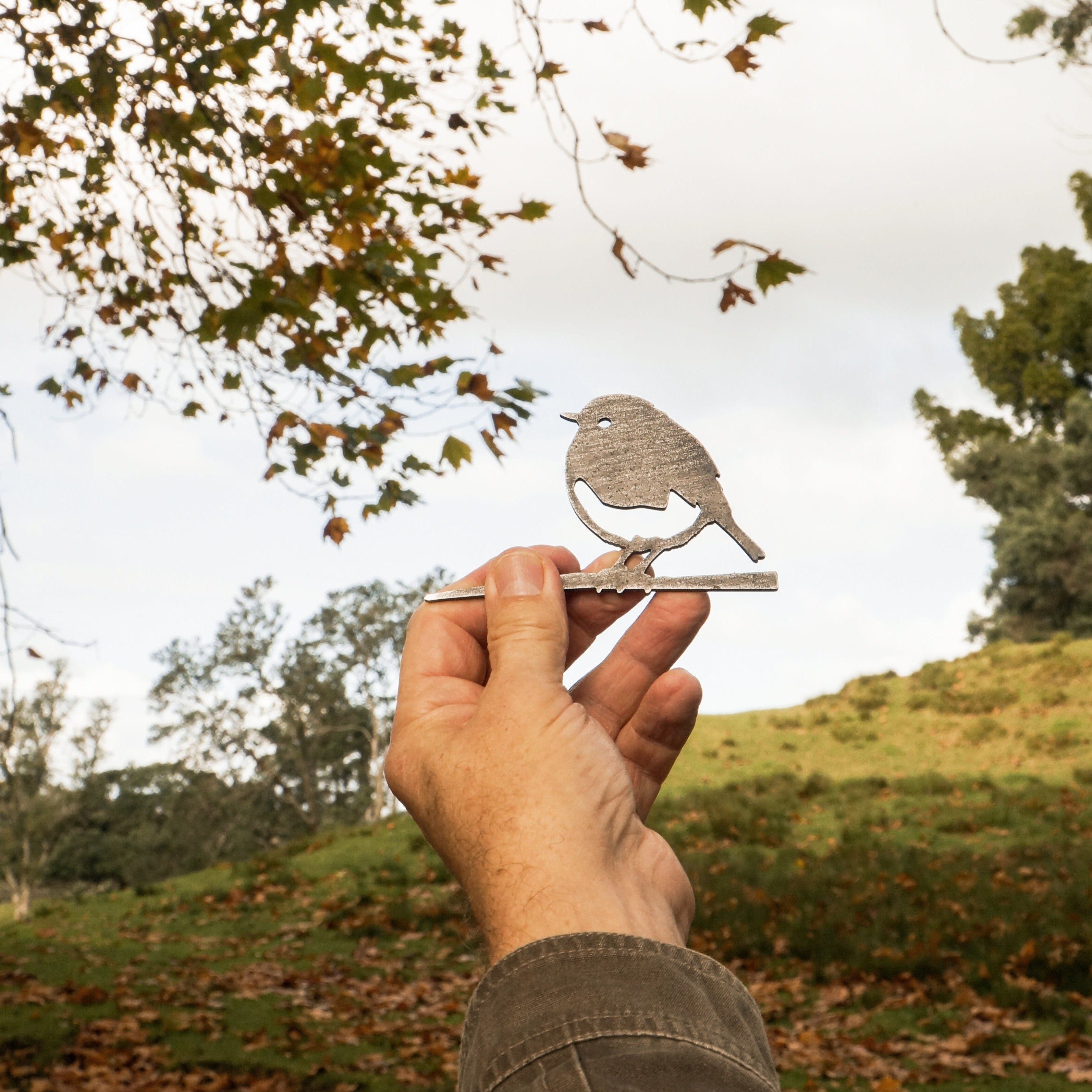 A hand holds up the Mini Robin by Metalbird UK outdoors, with trees, grass, leaves, and a cloudy sky in the background—an ideal gift for nature lovers.