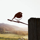 The Metalbird UK 'Robin' is a corten steel silhouette of a small bird on a branch, mounted on a wooden post, adding an elegant touch of garden art with a scenic sunset backdrop of green fields and hills.
