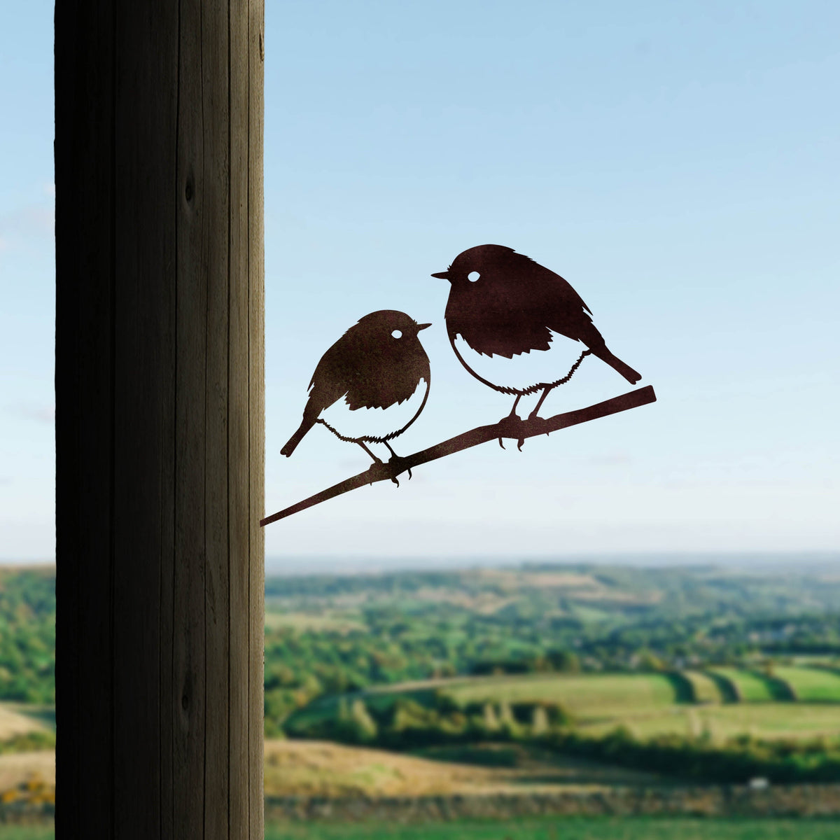Two steel Robin silhouettes on one branch installed in a wooden post overlooking fields.
