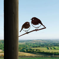 The Robin Pair by Metalbird UK features a corten steel silhouette of two robins on a branch, mounted on a wooden post with green fields and hills in the background—a charming gift for nature lovers.