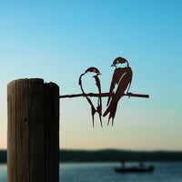 The Pair of Swallows by Metalbird UK features two metal bird silhouettes perched on a wire atop a wooden post, with a scenic lake and sunset sky backdrop—an ideal, charming gift for nature lovers.