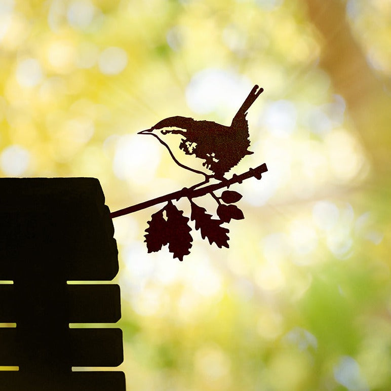 Steel Wren silhouette with an upright tail installed on a wooden surface against blurred foliage.
