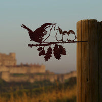 The Metalbird UK "Wren and Chicks" metal silhouette shows a bird feeding two chicks on a branch with oak leaves, mounted on a wooden post as striking garden art against a blurred landscape with a sunlit building in the distance.